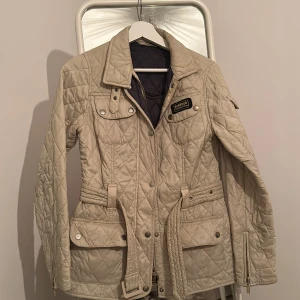 Beige quiltad jacka från Barbour - Säljer en snygg beige quiltad jacka från Barbour International. Jackan har ett bälte i midjan och flera fickor med knappar. Perfekt för höst och vår, med en stilren look och långärmad design. Passar både till vardags och lite finare tillfällen. Köpt ett tag sedan men har inte använts så mycket. Enstaka fläckar som kan försvinna med kemtvätt, 