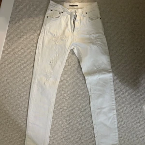 Vita jeans från Nudie Jeans - Säljer ett par vita jeans från Nudie Jeans i storlek 26/30. De har en snygg distressed look med slitningar på framsidan. De är i bra skick, förutom en litenliten fläck till vänster(på bild) om dragkedjan. 