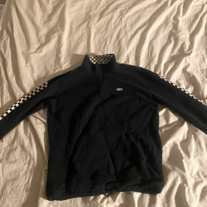 Vans zip-up - Säljer en svart tröja från Vans i bra skick. Den har en halv dragkedja och schackmönster längs ärmarna. Perfekt för en avslappnad stil och passar bra till både höst och vår. Tröjan har långa ärmar och är superbekväm! 
