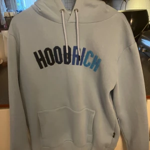Ljusblå hoodie från Hoodrich - Säljer en snygg ljusblå hoodie från Hoodrich i storlek L. Den har en stor ficka fram och text i olika blå nyanser på bröstet. Säljer även matchande byxor i storlek XL.