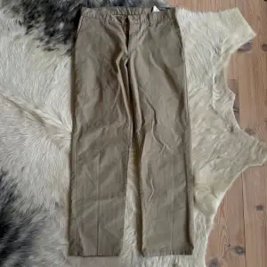 Säljer ett par klassiska beige byxor från Dickies i bra skick. De har en rak passform och är perfekta för både vardag och lite mer uppklädda tillfällen. Byxorna har en midwaist och är gjorda i ett slitstarkt material. Perfekta för höst och vår!