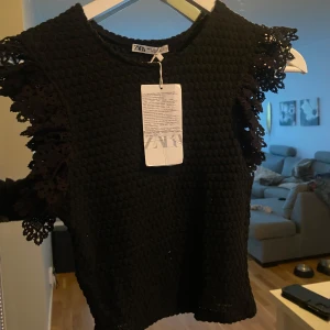 Svart topp från Zara - Snygg svart topp från Zara i ett stickat material med dekorativa spetsdetaljer på axlarna. Perfekt för både vardag och fest! Toppen är i nytt skick med etikett kvar.