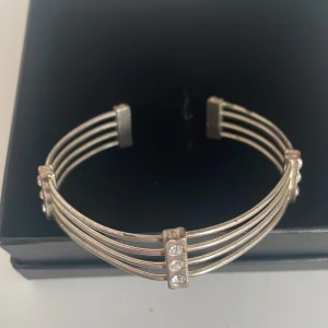 Silverfärgat armband med strassdetaljer - Snyggt och stilrent armband i silverfärg med flera parallella band och glittrande strassdetaljer. Perfekt för att ge en elegant touch till vilken outfit som helst. Armbandet har en öppen design som gör det enkelt att ta på och av.