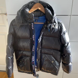 Svart pufferjacka från Polo Ralph Lauren - Säljer en snygg svart pufferjacka från Polo Ralph Lauren i storlek M i barn. Jackan har en glansig finish och är perfekt för kalla dagar. Den har en dragkedja och knappar framtill, samt en huva för extra värme. Insidan är fodrad med blått tyg och märket syns tydligt på bröstet. Perfekt för vintern! ❄️