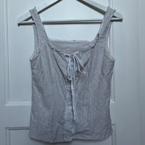 Vit ärmlös topp med knytning - Säljer en söt vit ärmlös topp med broderade blommor och knytning fram. Perfekt för sommardagar eller en avslappnad vårlook. Toppen har en fin passform och är i mycket bra skick.