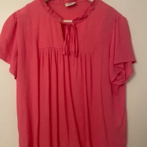 Rosa blus med knytband - Säljer en söt rosa blus med korta ärmar och knytband vid halsen. Blusen har en lätt rynkad design och är perfekt för vår och sommar. Materialet känns mjukt och luftig. Från Vero Moda