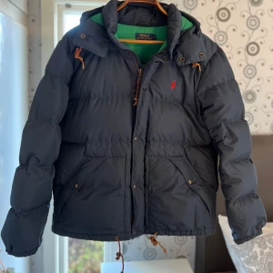 Svart pufferjacka från Polo Ralph Lauren - Säljer en snygg svart pufferjacka från Polo Ralph Lauren i storlek L. Jackan har en grön insida och är perfekt för kalla dagar. Den har en dragkedja och knappar framtill samt praktiska fickor. Perfekt för vintern! Storleken L/M