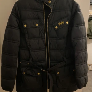 Svart quiltad jacka från Barbour - Säljer en svart quiltad jacka från Barbour. Den har en dragkedja och knappar framtill samt ett bälte i midjan för en justerbar passform. Perfekt för höst och vinter med långa ärmar och varma material. Jackan har flera fickor med knappar.