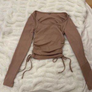 Beige ribbad topp med knyt - Säljer en superfin beige ribbad topp med långa ärmar och knytning i midjan. Perfekt för både vardag och fest! Den har en snygg, tight passform och är gjord i ett mjukt och stretchigt material. Toppen är i mycket bra skick och redo för en ny garderob.