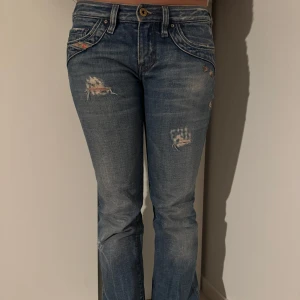 Blå jeans med slitningar - Säljer ett par snygga blå jeans från Diesel i bra skick. De har coola slitningar på framsidan och en klassisk femficksdesign. Jeansen är lågmidjade och har en straight passform, perfekt för en avslappnad stil. Passar både till vardags och fest! Midjemåttet är 38cm tvärs över, innebenslängd är 80cm 
