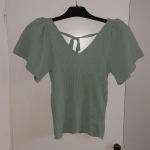 Grön ribbad topp med knyt - Säljer en superfin grön ribbad topp med korta ärmar och en söt knytning i ryggen. Perfekt för vår och sommar! Toppen har en v-ringad design som ger en elegant touch. Passar både till vardags och festliga tillfällen.
