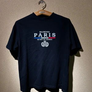 Svart t-shirt från Bal - Ej äkta snygg svart t-shirt med Bal tryck 'PARIS 40 RUE DE SEVRES' i vitt, blått och rött på framsidan. Perfekt för en casual look eller att klä upp med en kavaj. Kortärmad och i mjukt material för bekväm passform.