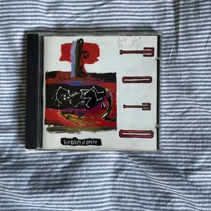 Kingdom of Desire - CD-skiva av bandet Toto med albumet 'Kingdom of Desire'. Omslaget har en abstrakt design i rött, svart och vitt. Skivan är i ett standard CD-fodral med en transparent framsida och svart baksida. Perfekt för samlare eller fans av Toto!