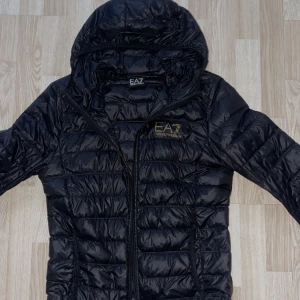 Svart pufferjacka från Emporio Armani - Säljer en EA7 jacka, använt den ett tag och finns ett litet hål i ryggen som inte syns så mycket men måste ändå peka ut det och anledningen varför jag säljer den är för att den har blivit för liten för mig!