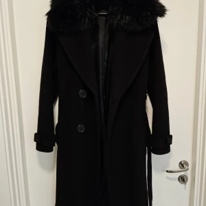 EKOL svart kappa, storlek S - Svart kappa med pälskrage. Kappan har en midjeband och fyra knappar framtill.  Storlek 36-38/S.
