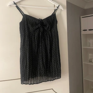 Svart prickig linne - Säljer ett svart linne med vita prickar. Det har smala axelband och en söt knytdetalj framtill. 