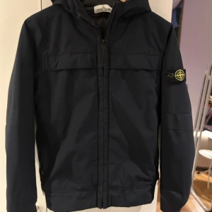 Svart jacka från Stone Island - Stone Island jacka. Junior strl 12/156 Mycket bra skick. Jackan har en dragkedja framtill och huva. Ej avtagbar. 