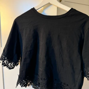 Svart topp med spetsdetaljer - Säljer en snygg svart topp med korta ärmar och vackra spetsdetaljer vid ärmslut och nederkant. Perfekt för en stilren och chic look. Toppen är croppad och passar bra till både vardag och fest.