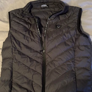 Svart dunväst från Emporio Armani - Snygg svart dunväst från Emporio Armani med dragkedja och logotypdetalj på bröstet. Perfekt för höst och vår, håller dig varm och stilren. Västen har en quiltad design och är i mycket bra skick. Passar både till vardags och lite finare tillfällen.