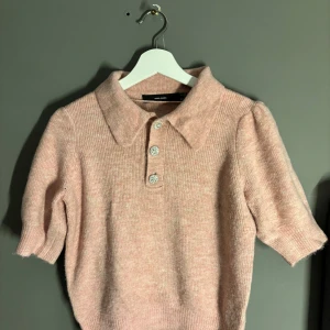 Rosa stickad tröja från Vero Moda - Superfin rosa stickad tröja från Vero Moda i storlek S. Den har en klassisk krage och tre dekorativa knappar framtill. Perfekt för både vår och höst med sina korta ärmar och mjuka material. En mysig och stilren tröja för alla tillfällen!