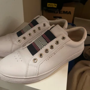 Vita sneakers från Tommy Hilfiger - Säljer ett par stilrena vita sneakers från Tommy Hilfiger. De har en cool elastisk rem med mönster i svart och rött istället för traditionell snörning. Perfekta för en avslappnad men ändå snygg look. Använda men i mycket bra skick.