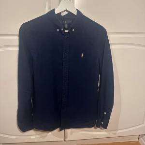 Mörkblå skjorta från Ralph Lauren - Säljer en snygg mörkblå skjorta från Ralph Lauren i mycket bra skick. Skjortan har långa ärmar och klassiska knappar framtill. Den är perfekt för både vardag och fest. Den ikoniska loggan är broderad på bröstet. 