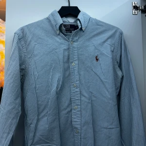 Randig skjorta från Polo Ralph Lauren - Säljer en snygg och klassisk randig skjorta från Polo Ralph Lauren i storlek S. Skjortan är blå och vit med långa ärmar och knappar framtill. Perfekt för både vardag och festliga tillfällen. Den har en broderad logga på bröstet som ger en extra touch av stil. Passar perfekt till jeans eller chinos!