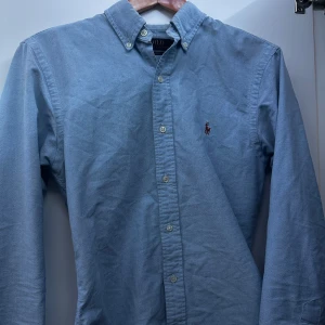 Blå skjorta från Polo Ralph Lauren - Säljer en snygg blå skjorta från Polo Ralph Lauren i storlek S. Skjortan har en klassisk design med knappar och en liten broderad logga på bröstet. Den är i slim fit och passar perfekt för både vardag och fest. Skjortan är långärmad och gjord i ett bekvämt material.