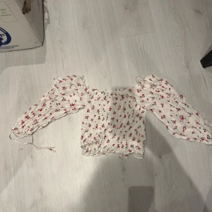Blommig offshoulder-top - Säljer en superfin blommig offshoulder-top i vitt med små röda blommor. Toppen har långa ärmar och en söt volangdetalj vid ärmslut och nederkant. Perfekt för vår och sommar! 🌸