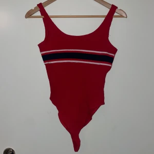 Röd body med randig detalj - Snygg röd body med breda axelband och en cool randig detalj i marinblått och vitt över bröstet. Låg i ryggen men ej helt öppen. Aldrig använd men haft i min garderob väldigt länge. Storlek M men passar bättre en S.