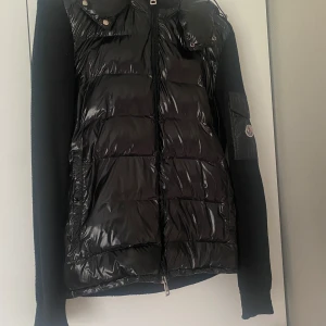Svart pufferjacka från Moncler - Säljer en snygg svart pufferjacka från Moncler. Jackan har en glansig finish med dragkedja framtill och långa ärmar. Perfekt för kyliga dagar när du vill hålla stilen. Moncler-loggan syns på ärmen. Passar både till vardags och lite finare tillfällen.
