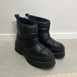 Svarta vadderade boots Pavement strl 38 - Säljer ett par svarta vadderade boots i mycket bra skick. De har en chunky sula och är perfekta för vintern. Bootsens insida är fodrad för extra värme och komfort. De är lätta att ta på tack vare de praktiska flikarna baktill. Perfekta för att hålla fötterna varma och stiliga under kalla dagar! Knappt använda. Säljer pga. att jag använder mina andra vinterskor mer.