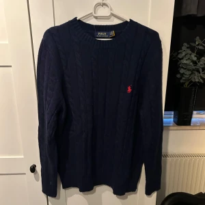 Mörkblå stickad tröja från Polo Ralph Lauren - Säljer en snygg mörkblå stickad tröja från Polo Ralph Lauren. Den har en klassisk design med det ikoniska röda logotypbroderiet på bröstet. Perfekt för både höst och vinter, och den är i nytt skick. Passar till både vardag och lite finare tillfällen.