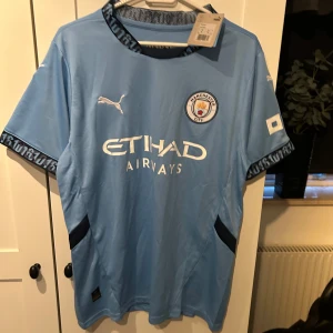Ljusblå Manchester City fotbollströja - Säljer en ljusblå Manchester City tröja från Puma i nytt skick. Den är kortärmad med snygga detaljer på kragen och ärmsluten. Perfekt för alla City-fans! Tröjan har klubbens emblem och sponsorlogga på framsidan. Materialet är lätt och andas bra, vilket gör den idealisk för både träning och match.