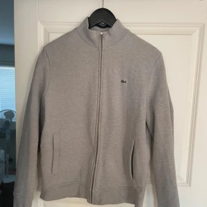 Grå kofta från Lacoste - Säljer en stilren grå jacka från Lacoste i mycket bra skick. normal passform. Koftan är gjord i ett mjukt bomullsmaterial och Passar både till vardags och lite finare tillfällen.