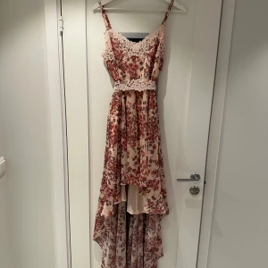 Blommig klänning med spetsdetaljer - Superfin blommig klänning med spetsdetaljer och asymmetrisk längd. Den har smala axelband och en vacker spetskant vid halsen. Perfekt för sommarens fester eller en romantisk kväll. Klänningen har en ljus bas med röda blommor och en elegant passform.