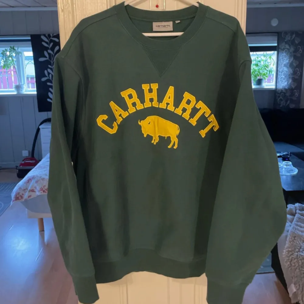 Säljer en snygg grön sweatshirt från Carhartt med gult tryck av en buffel och texten 'CARHARTT' på framsidan. Tröjan är i mycket bra skick och perfekt för både vardag och chill. Den har en normal passform och är superbekväm. Perfekt för höst och vår!. Tröjor & Koftor.