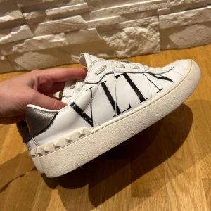 Valentino Garavani sneakers i vitt och silver - Säljer ett par snygga Valentino Garavani sneakers i vitt med svarta detaljer och silver på hälen. Skorna har en cool design med VLTN-logga och nitar runt sulan. Perfekta för att ge din outfit en lyxig touch! Använda men i bra skick.
