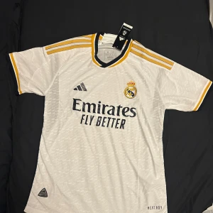Vit Real Madrid fotbollströja - Säljer en ny vit Real Madrid fotbollströja från Adidas. Den har korta ärmar och snygga gula detaljer på axlarna. Tröjan är i HEAT.RDY-material för optimal komfort. Perfekt för alla Madridistas! 💛⚽