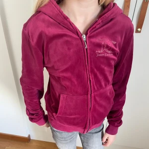 Vinröd hoodie från Juicy Couture - Säljer en supermjuk och snygg vinröd hoodie från Juicy Couture. Den har en dragkedja framtill och en stor luva. På ryggen finns det ett glittrigt Juicy Couture-tryck. Perfekt för en chill dag eller en avslappnad utflykt. Passar perfekt till jeans eller leggings!