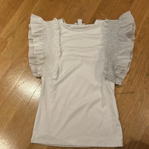 Vit topp med volangärmar från Amisu - Säljer en superfin vit topp från Amisu i storlek S. Den har korta ärmar med volangdetaljer. Perfekt för både vardag och fest! Toppen är i mycket bra skick och redo för en ny ägare.
