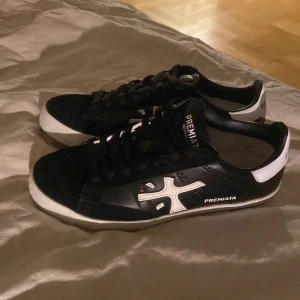 Premiata skor - Snygga svarta sneakers från Premiata i mycket bra skick. De har en stilren design med vita detaljer och logga på sidan. Skorna har en bekväm passform och är perfekta för både vardag och fest. Sulan är vit med subtila mönster och skorna har klassisk snörning. Nypris runt 3000kr