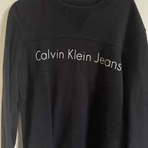  Calvin Klein Jeans Sweatshirt  - Säljer en stilren svart tröja från Calvin Klein Jeans. Den har en enkel design med märkets logga tryckt över bröstet. Perfekt för en casual look och passar till det mesta. Tröjan är i mycket bra skick och har långa ärmar.