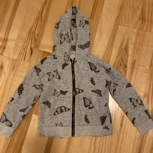 Grå hoodie med fjärilsmönster från Name It - Säljer en superfin grå hoodie från Name It med ett gulligt fjärilsmönster. Den har en dragkedja framtill och en mysig huva. Perfekt för både vår och höst när det är lite kyligt ute. Passar barn i storlek 122-128.