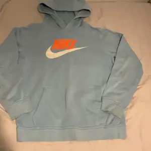 Säljer en snygg ljusblå hoodie från Nike med den klassiska loggan i orange och vitt på framsidan. Den har en stor ficka fram och är perfekt för både chill och sportiga dagar. Superbekväm och stilren!