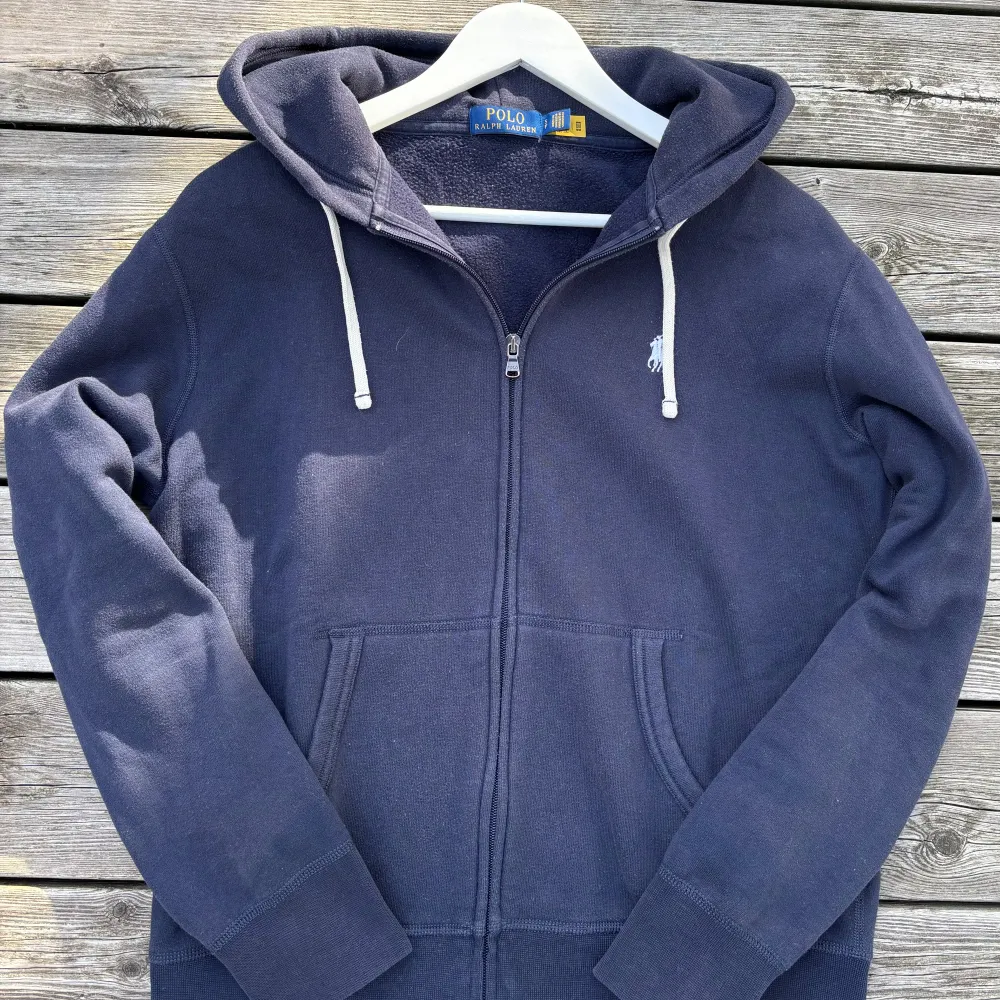 Säljer en snygg och bekväm mörkblå hoodie från Ralph Lauren. Den har en dragkedja framtill och en klassisk huva med vita snören. Perfekt för kyliga dagar och har en stilren design med det ikoniska Ralph Lauren-märket på bröstet. Passar både till vardags och lite mer avslappnade tillfällen.. Tröjor & Koftor.