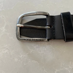 Svart skärp från Jack & Jones - Säljer ett stilrent svart skärp från Jack & Jones. Det är gjort av skinn och har ett klassiskt silverfärgat spänne med märkets logga ingraverad. Perfekt för både vardag och fest! 🖤