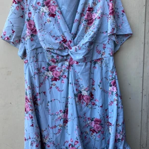 Blommig klänning i ljusblått - Säljer en superfin ljusblå klänning med blommigt mönster i rosa och vinrött. Klänningen har en v-ringad omlottdesign framtill och korta ärmar. Perfekt för sommarens fester eller en dag på stan!