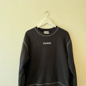 Svart sweatshirt från Ganni - Säljer en svart sweatshirt från Ganni i storlek M. Den har ett coolt tryck med text och en blomma på baksidan. Tröjan är långärmad och har vita sömmar som ger en snygg kontrast. Perfekt för en avslappnad stil!