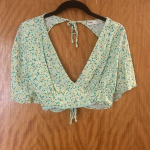 Blommig croppad topp med knytband - Säljer en superfin blommig croppad topp från pull & bear. Den har en djup v-ringning och öppen rygg med knytband. Toppen är i en ljusgrön nyans med små blommor i blått och gult. Perfekt för solsemester eller en fest kväll!